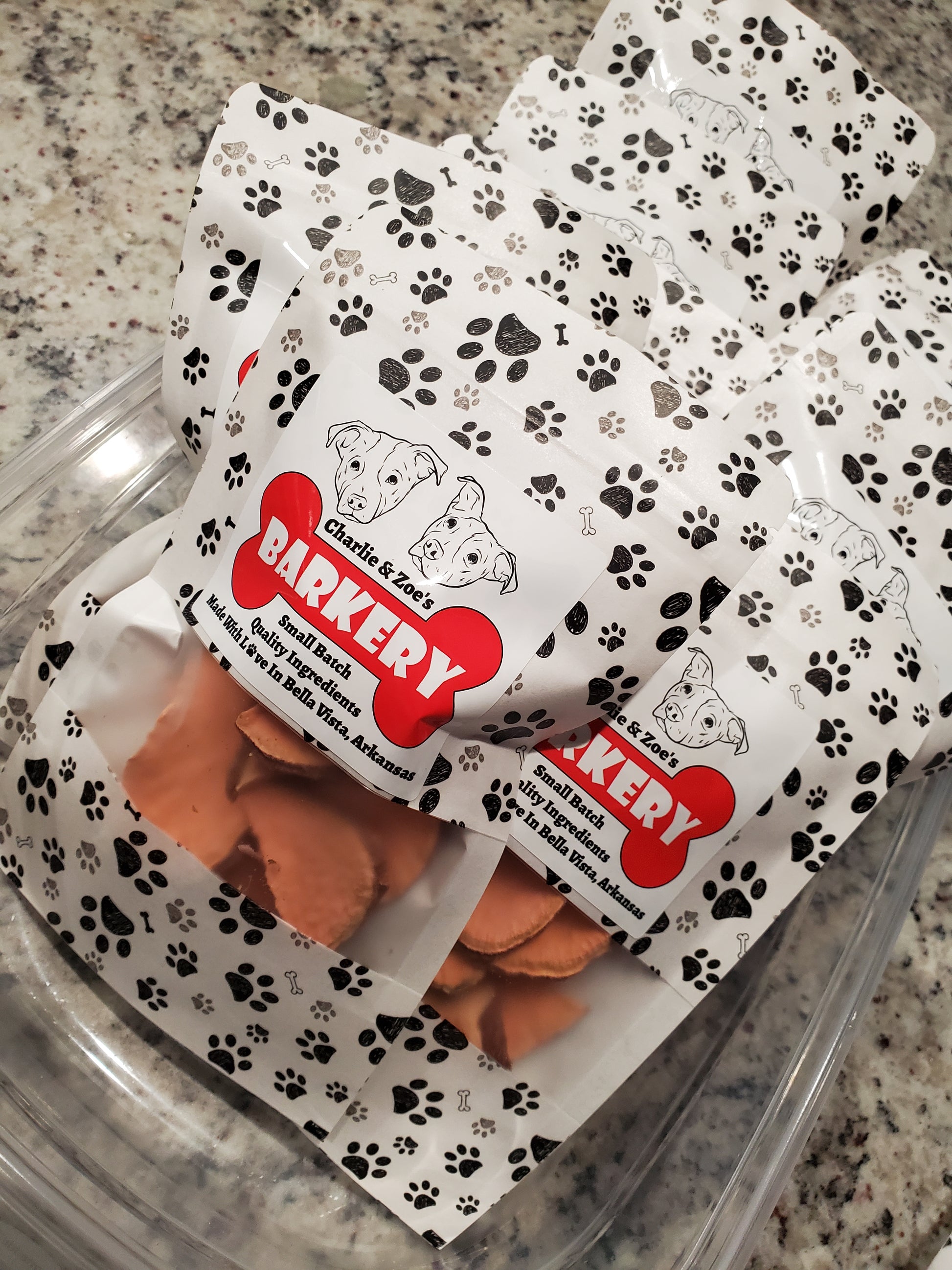 Dog treats in a clear container with Charlie & Zoe's Barkery packaging on a granite surface